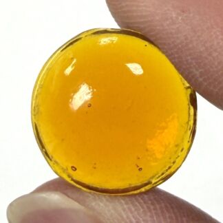 Circular amber gemstone held between two fingertips against a white background with light reflecting off its surface.