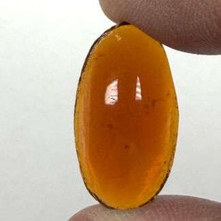 Bright orange oval gemstone held between thumb and index finger against a pale background, showing a smooth, translucent surface outside light glare.